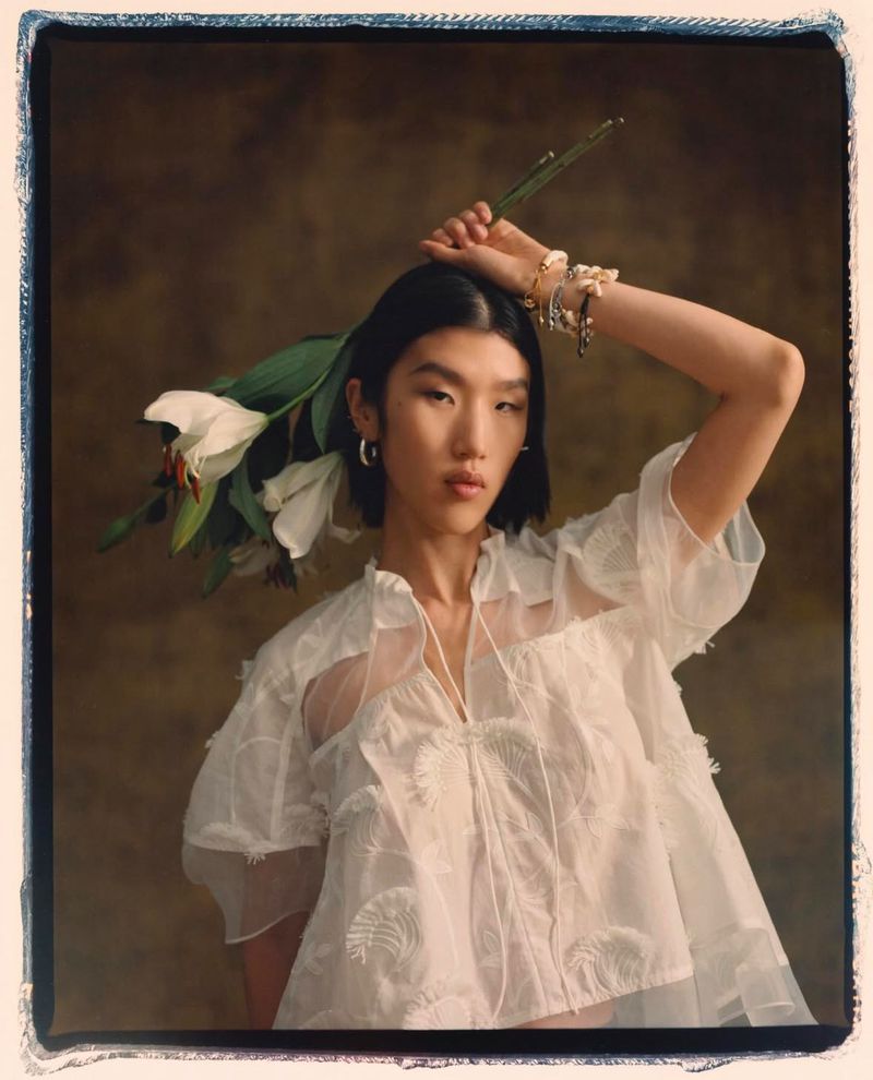 Young Asian woman poses in a sheer white blouse, holding a white lily against a blurred olive green background in a fashion editorial.
