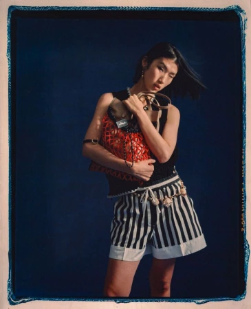 Model holding a red mesh bag and wearing striped shorts against a dark blue textured studio background.