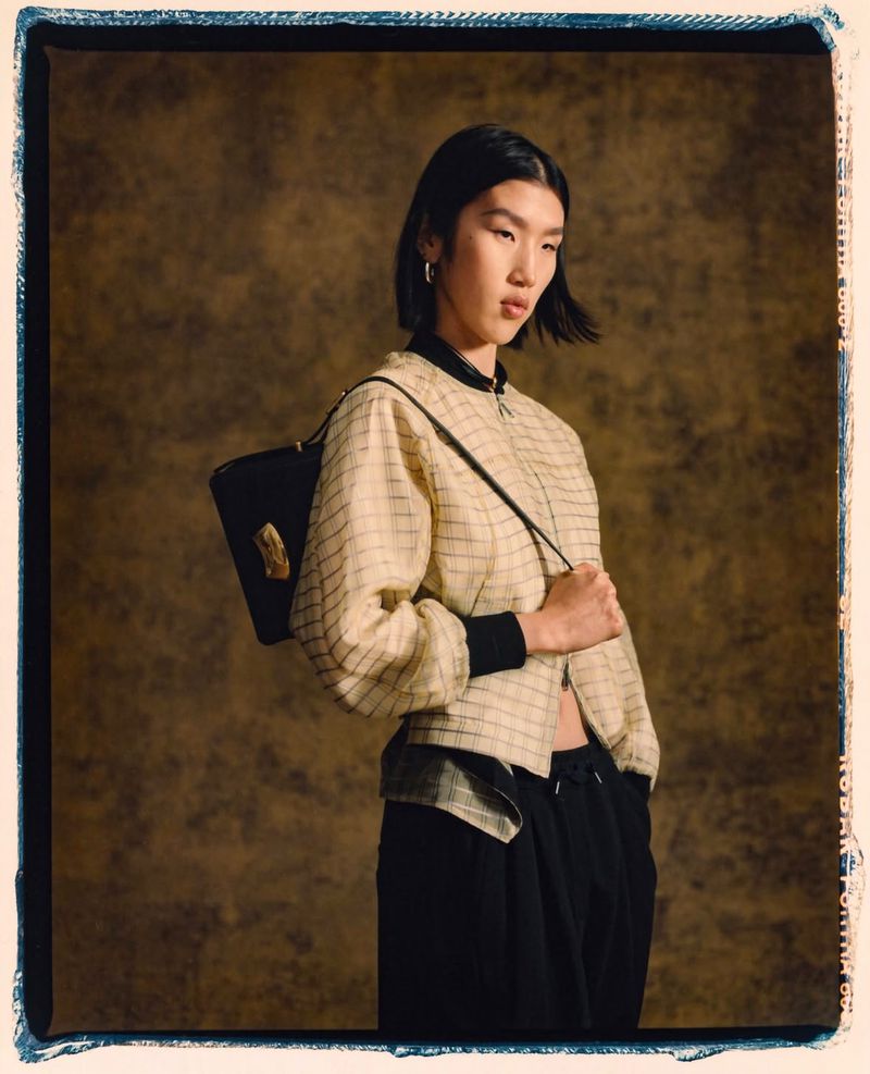 Asian woman in a checkered jacket stands in front of a mottled brown backdrop, holding a black purse over her shoulder in studio lighting.