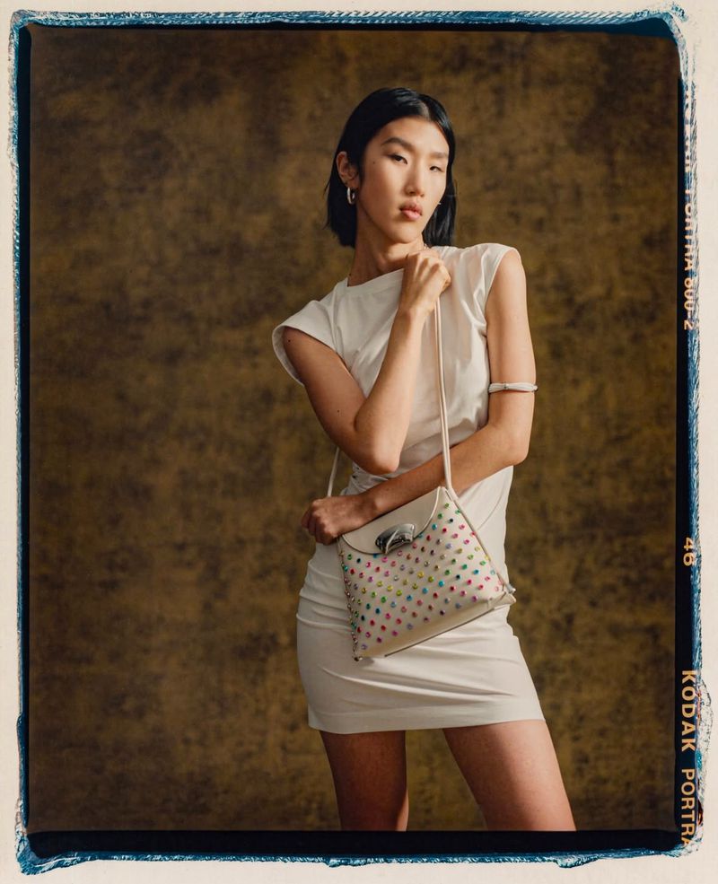 Asian woman in a short white dress poses in front of a textured brown backdrop, holding a handbag with colorful polka dot details.