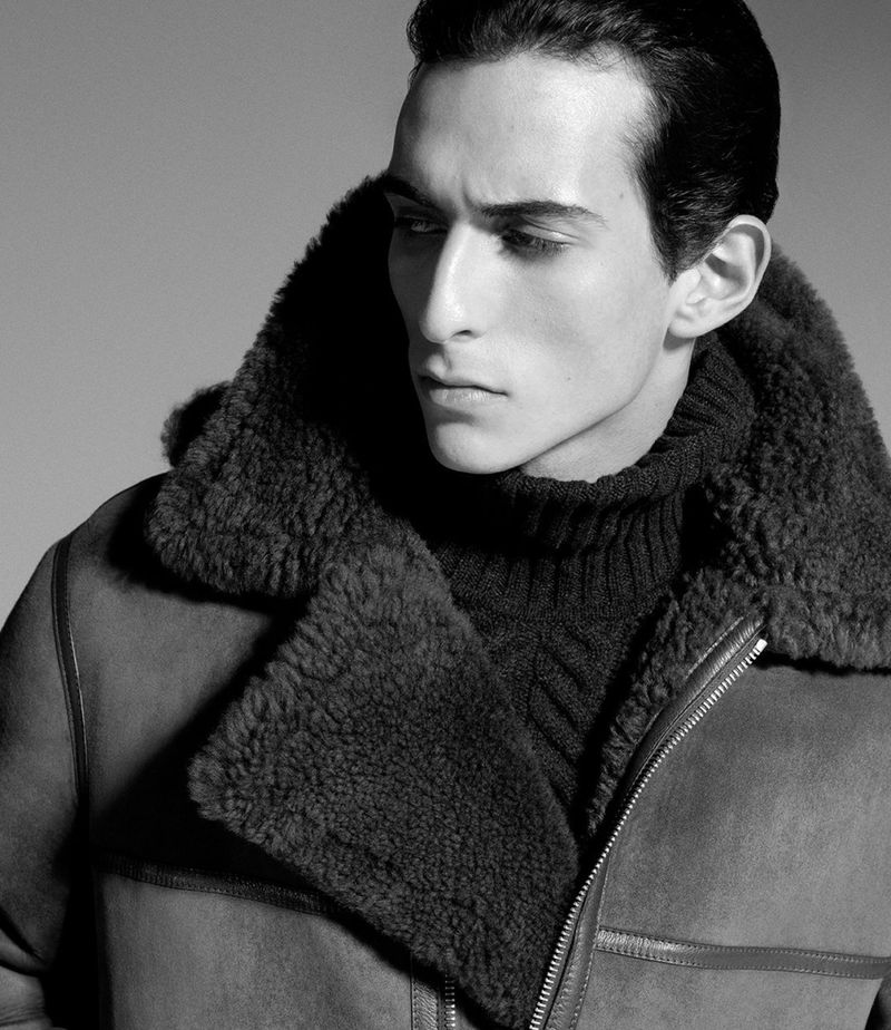 Monochrome portrait of a man in a shearling coat and turtleneck sweater, eyes looking off-camera, in a grey studio setting.