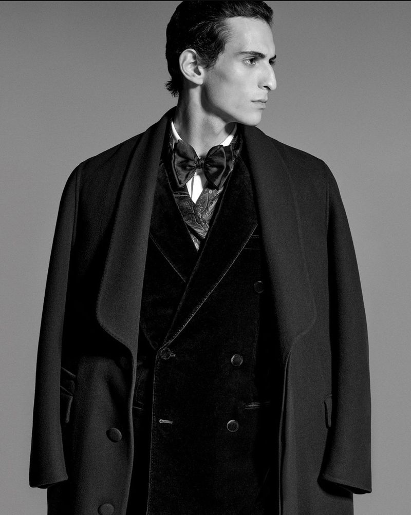Black-and-white portrait of a slender man in a velvet tuxedo vest, bow tie, and formal coat posing in a photo studio.