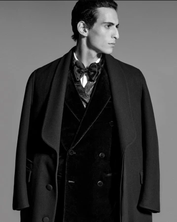 Black-and-white portrait of a slender man in a velvet tuxedo vest, bow tie, and formal coat posing in a photo studio.