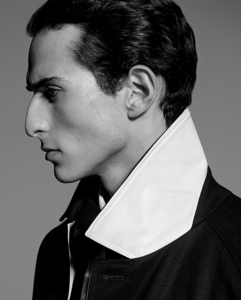 Monochrome portrait of a dark-haired man in a coat with a white collar, captured in a side view against a grey backdrop.