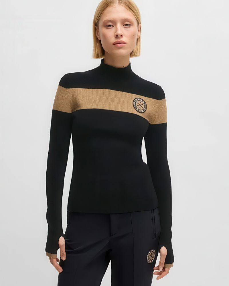 A fair-skinned young adult female posing indoors, wearing a black and tan long-sleeved knit top with black pants against a neutral background.