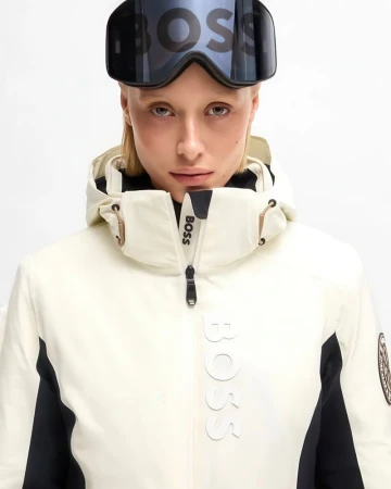 Close-up of a fair-skinned woman wearing black ski goggles and a white and black ski jacket with 'BOSS' logos against a white backdrop.