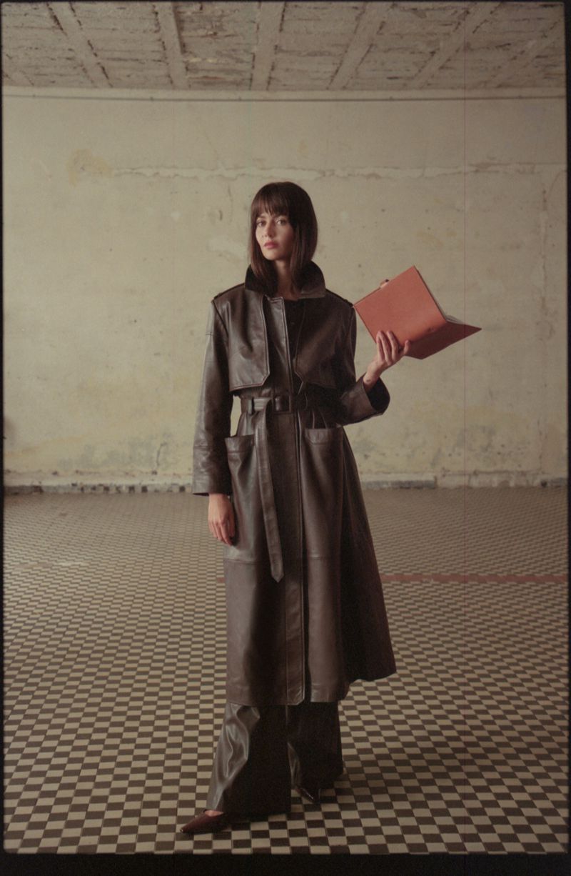 A stylish woman in a long brown leather coat stands in a tiled room, holding a laptop, blending fashion with technology indoors.
