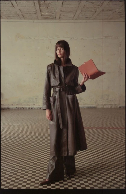 A stylish woman in a long brown leather coat stands in a tiled room, holding a laptop, blending fashion with technology indoors.