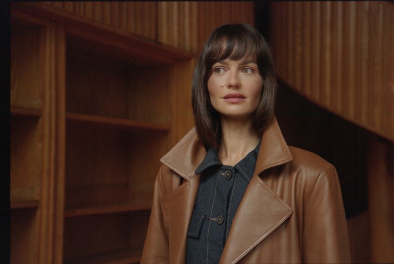 Portrait of a woman with brown hair and brown coat in front of a wooden wall and shelves in an indoor setting, looking off-camera.