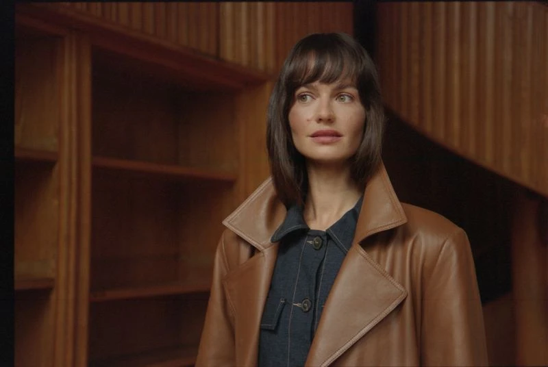 Portrait of a woman with brown hair and brown coat in front of a wooden wall and shelves in an indoor setting, looking off-camera.