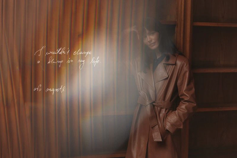 Woman in a brown leather trench coat stands next to a bookshelf with handwritten script overlaying the image, creating a vintage and artistic vibe.