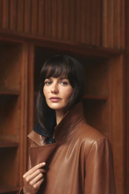 Portrait of a woman with brown hair wearing a tan leather trench coat, posing indoors against a dark wooden bookshelf background.