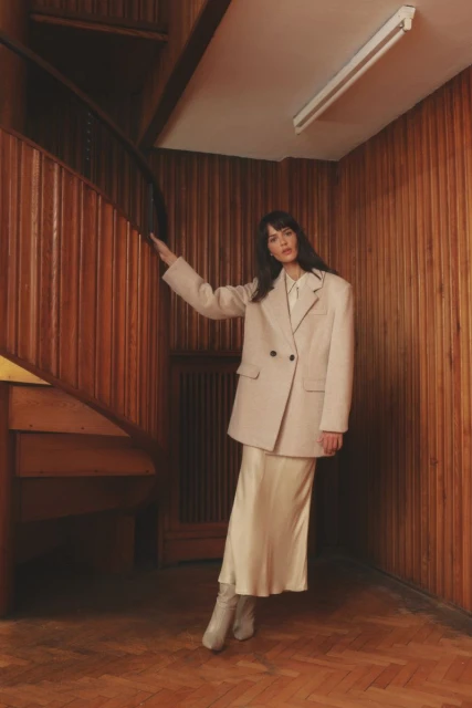 Brunette woman in cream-colored blazer and matching skirt and boots stands in a wood-paneled stairwell near a wooden staircase.