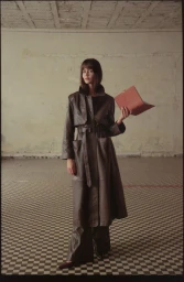 Model Marta Dyks wearing a long brown leather coat holding a notebook in an empty room photographed by Paulina Manterys
