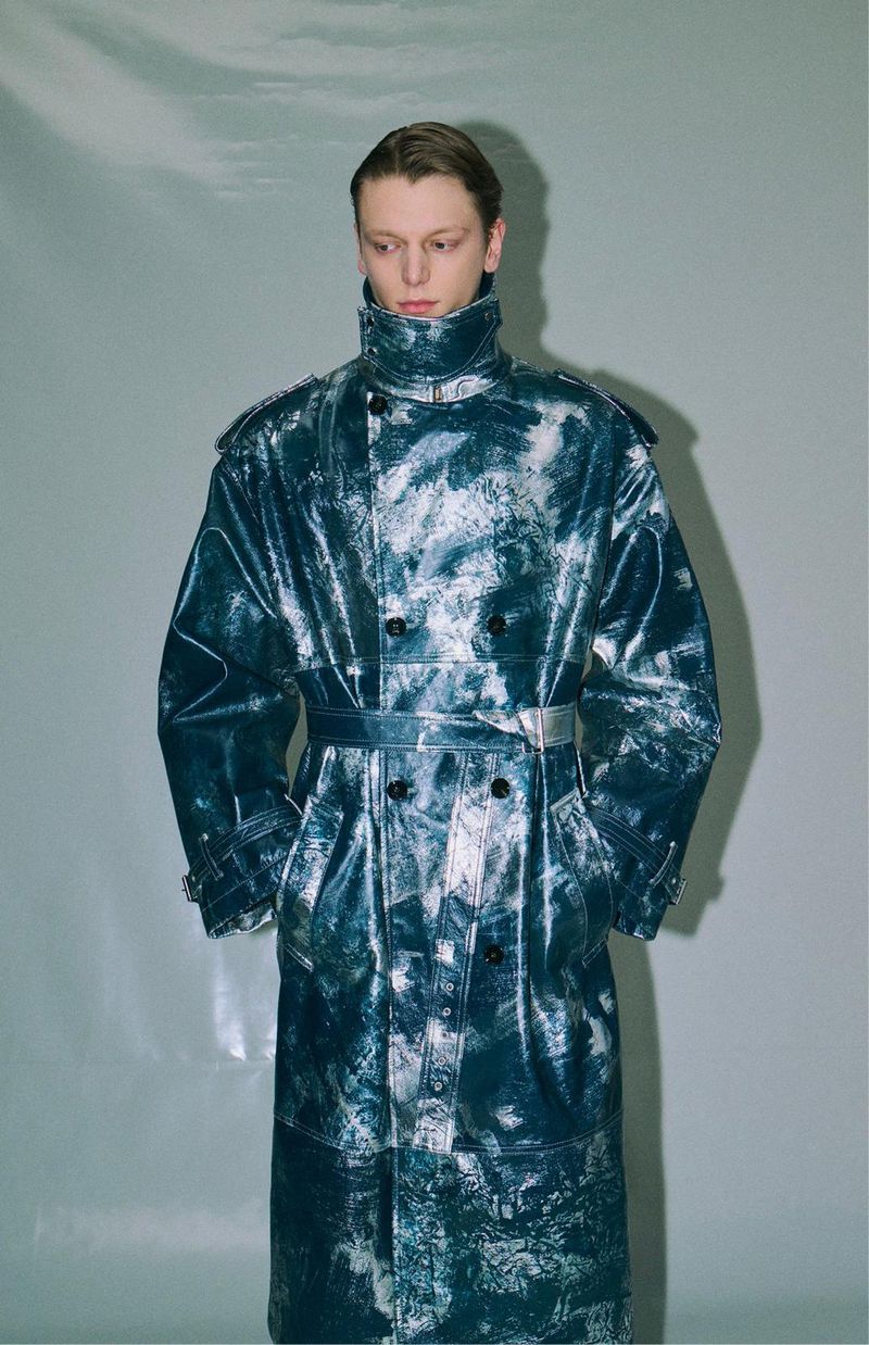 A person wearing a long, patterned, blue and white coat, standing indoors against a light gray wall with some reflections.