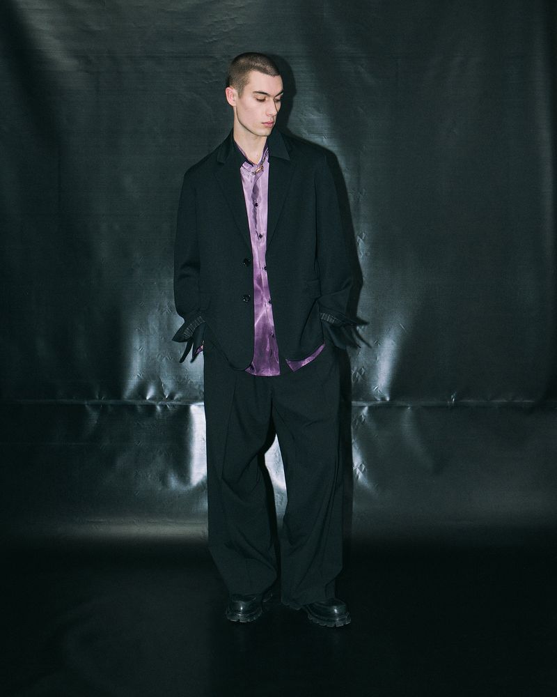 Man in black suit and purple shirt poses in a full shot against a dark background. Fashion shot with dramatic shadow and formal wear.