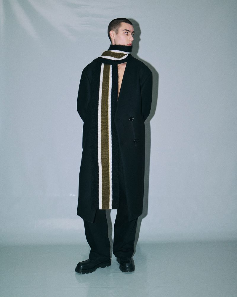 A slender man in a black coat and black pants stands indoors, wearing a striped scarf. He gazes off to the side in a fashion editorial shot.