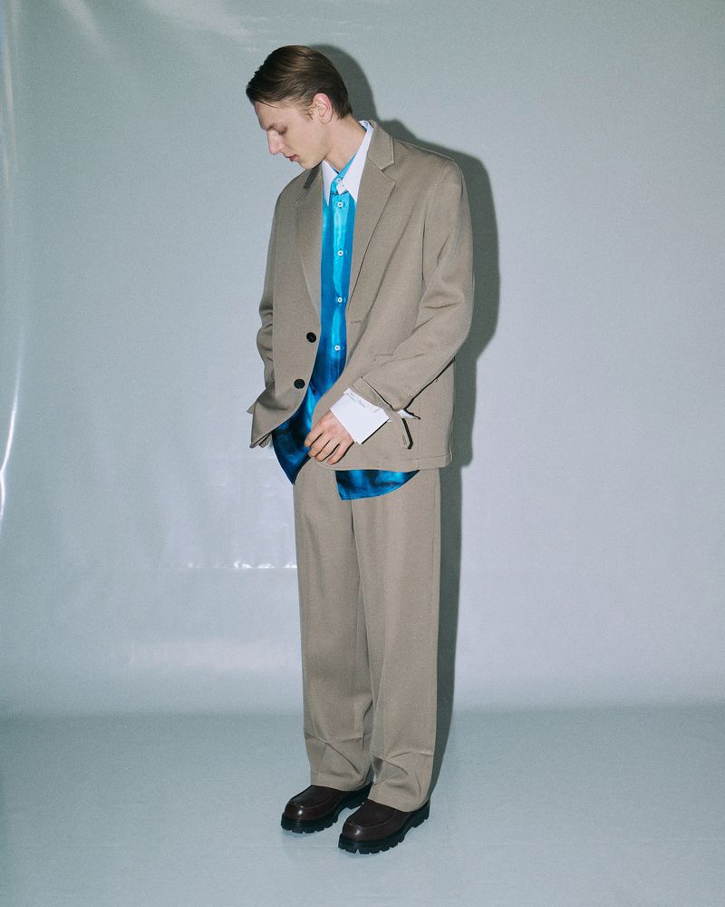 Full-length shot of a young adult male in a tan suit with a bright blue shirt, posing indoors against a gradient white background.