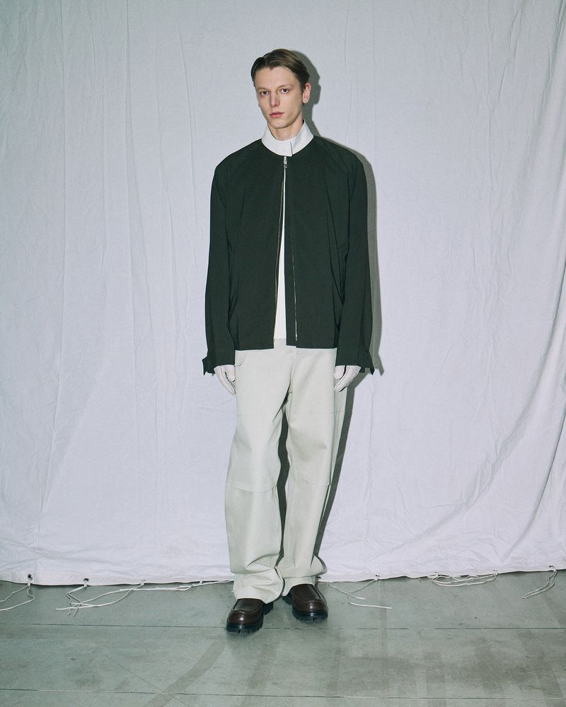 Male fashion model poses in an indoor photo studio wearing a dark green jacket, white collared shirt, white pants, and brown shoes.