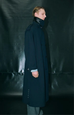 Profile view of a model in a long dark coat standing against a dark glossy textured background.