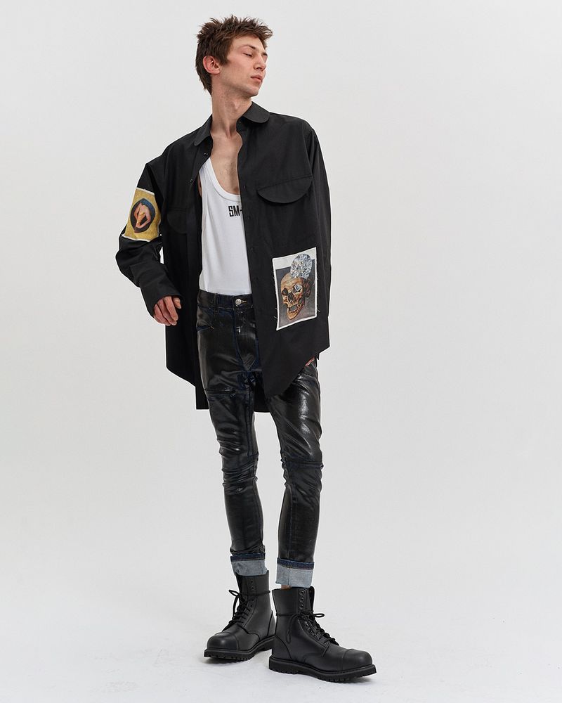 Young adult man in black shirt, white tank top, dark jeans and boots posing in a full shot against a white backdrop.