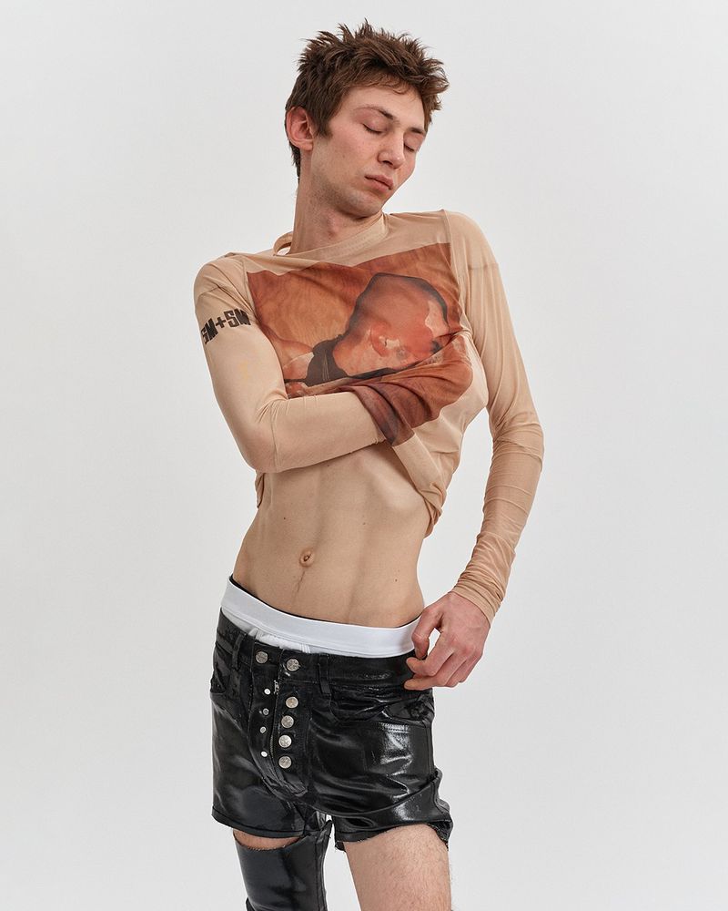 A slender adult male poses indoors wearing a sheer printed long-sleeved shirt and black shorts, set against a neutral background in a studio.