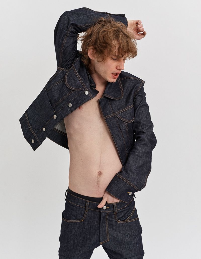 A young adult man poses in a denim jacket and jeans against a neutral backdrop, showcasing a fashion-forward, casual style.