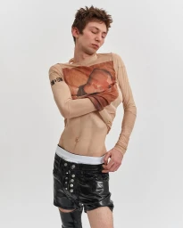 A male model wearing a sheer graphic long-sleeve top and black shiny shorts against a plain white background.