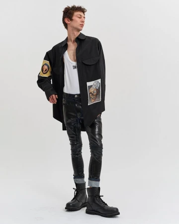 Model wearing a black shirt with patches over a white tank top and dark trousers against a white backdrop.