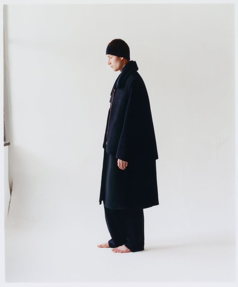 A person in a dark robe and head covering stands sideways in a brightly lit white studio, showing fashion and style elements.