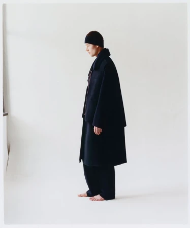 A person in a dark robe and head covering stands sideways in a brightly lit white studio, showing fashion and style elements.