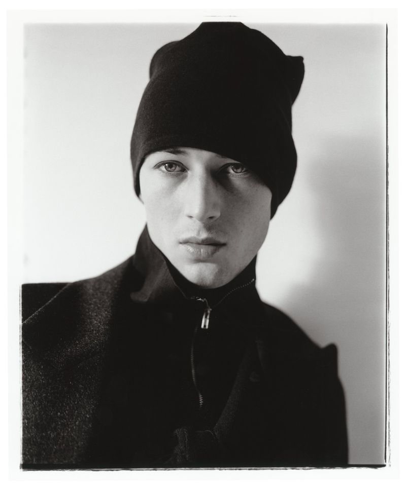 Monochrome portrait of a young adult male in a dark coat and beanie against a plain background, capturing a contemplative and artistic mood.