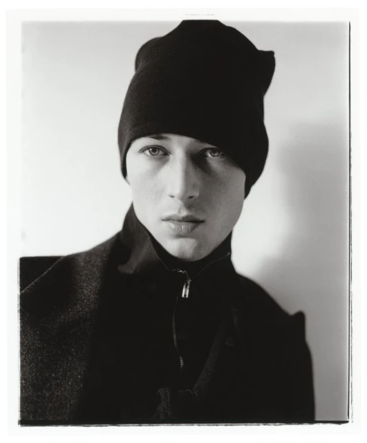 Monochrome portrait of a young adult male in a dark coat and beanie against a plain background, capturing a contemplative and artistic mood.