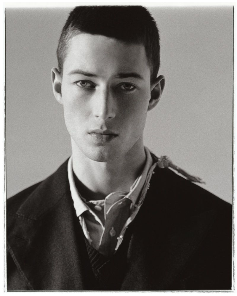 Monochrome portrait of a young adult male model in a dark blazer, unbuttoned collared shirt, and patterned neckerchief against a grey backdrop.