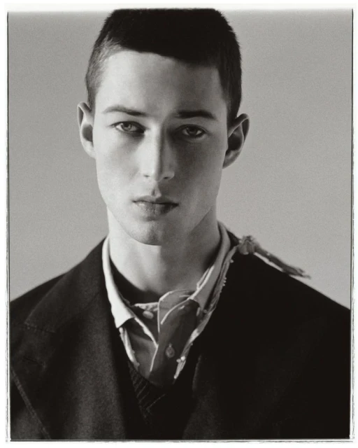 Monochrome portrait of a young adult male model in a dark blazer, unbuttoned collared shirt, and patterned neckerchief against a grey backdrop.