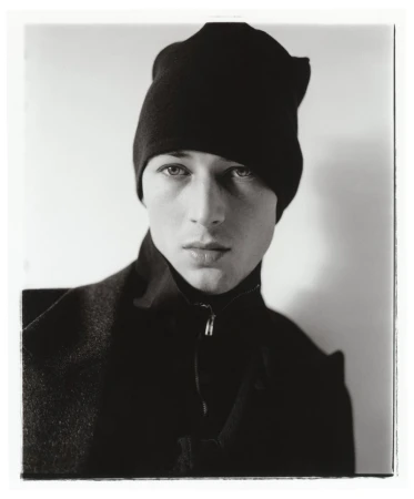 Portrait of a male model wearing a black beanie and black zipped jacket against a plain background.