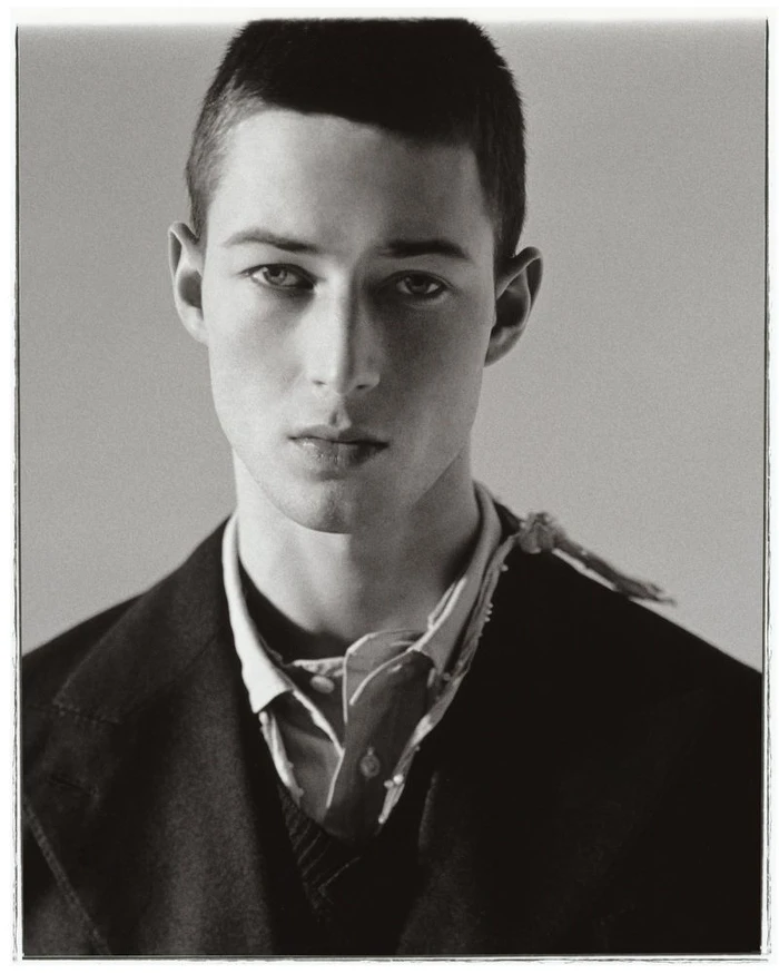 Portrait of a young male model with short hair wearing a dark jacket and collared shirt against a plain grey studio background.