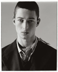 Portrait of a young male model with short hair wearing a dark jacket and collared shirt against a plain grey studio background.