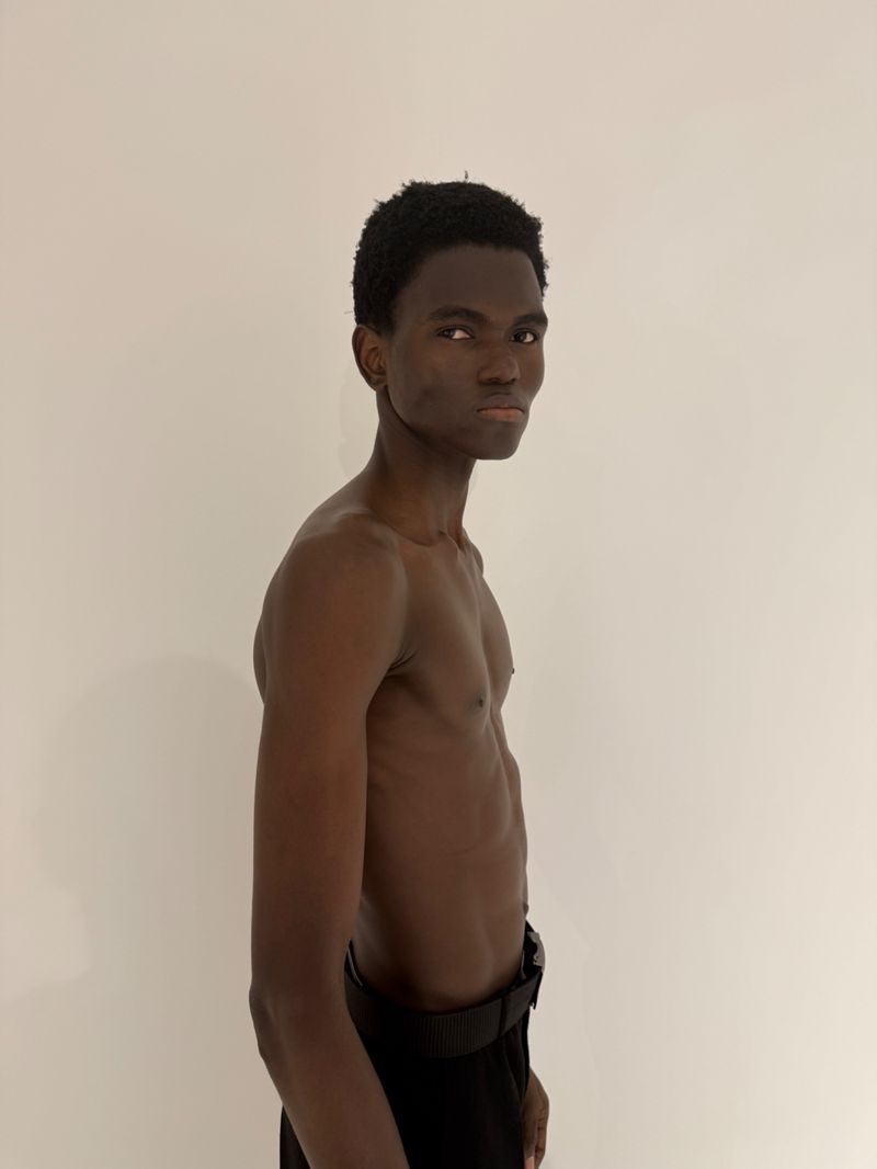 A shirtless young Black man stands in profile, looking over his shoulder against a plain white background.