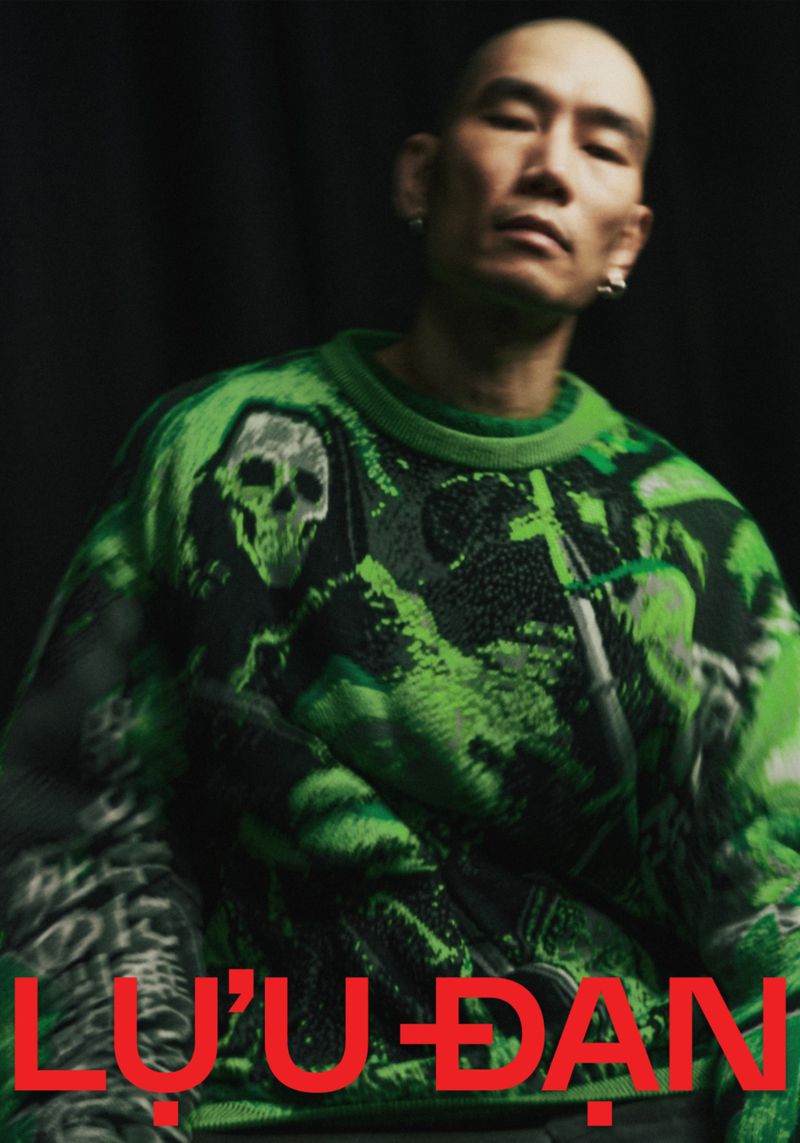 Adult male with shaved head poses in a dark environment wearing a patterned green and black sweater with text at the bottom.