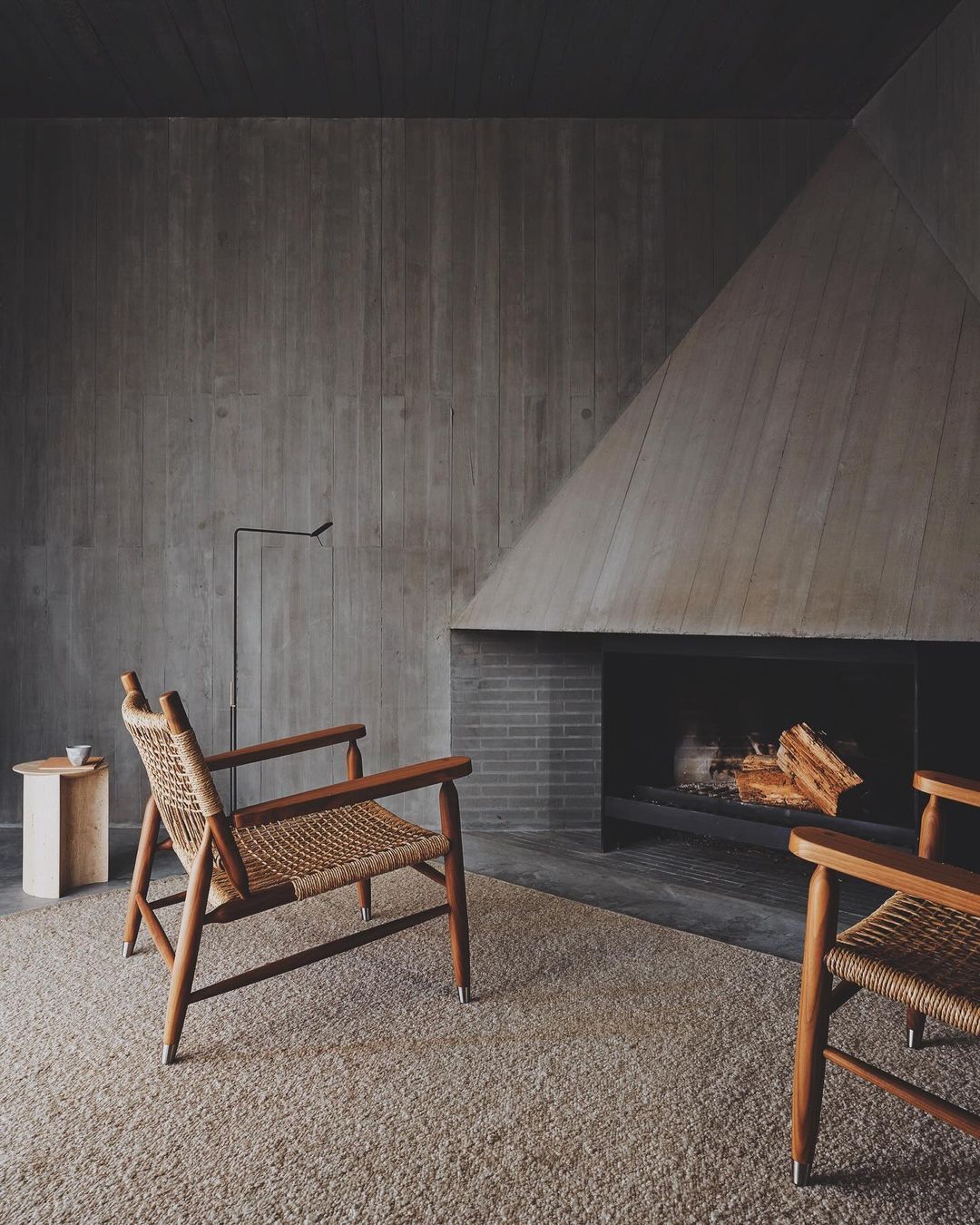 Interior design featuring two wooden armchairs, a fireplace, wooden side table, and a minimalist lamp, set against a concrete wall.