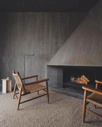 Two wooden armchairs with woven seats rest on a textured rug in a minimalist living room with a concrete fireplace.