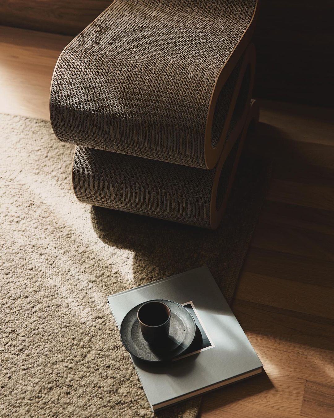 Stacked cardboard chair near a cup on a plate and book on a rug, placed on hardwood flooring near a wall.