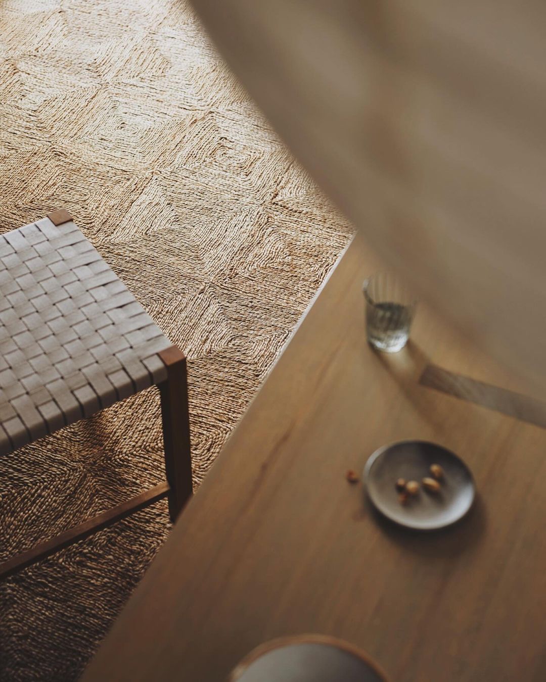 Interior design featuring a woven rug, wooden chair, and table with a glass of water and dish with nuts, showcasing a neutral color palette.
