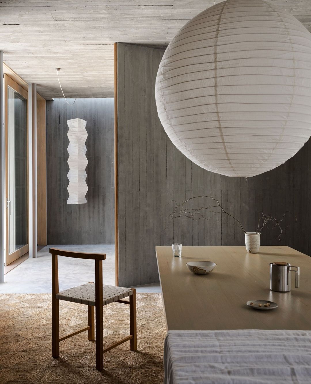 Interior view of a dining area with a minimalist wooden chair, a table set with tableware, and contemporary paper lanterns.
