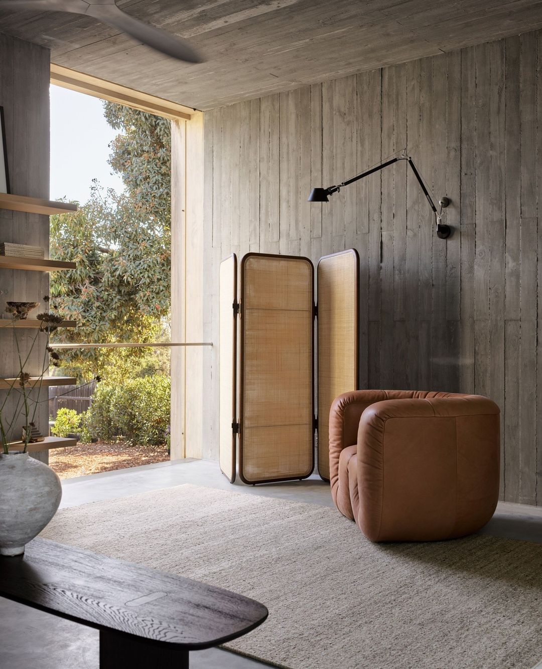 Interior of a modern living space featuring a tan leather chair, a folding screen, and a concrete wall illuminated by natural light.