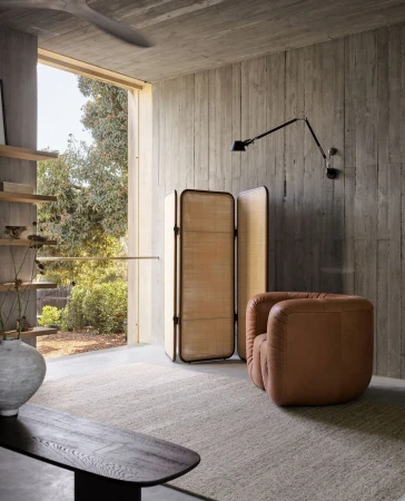 Interior of a modern living space featuring a tan leather chair, a folding screen, and a concrete wall illuminated by natural light.