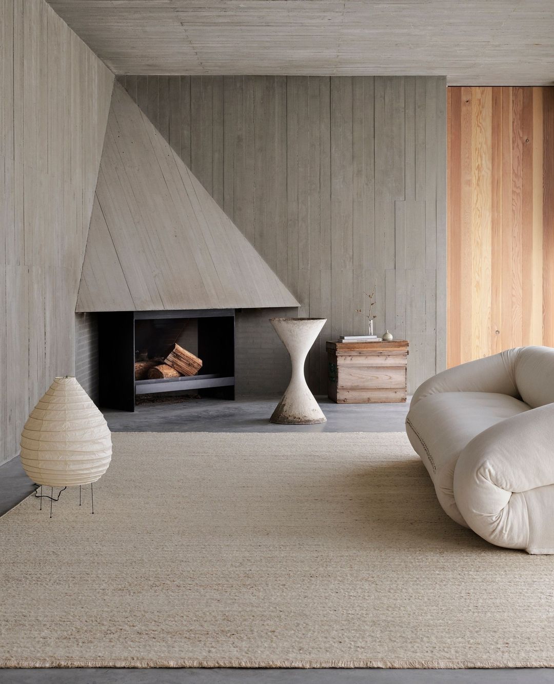 Minimalist interior featuring a tan rug, modern couch, black fireplace, and unique hourglass sculpture against a concrete wall.
