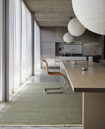 Minimalist interior featuring a modern chair, neutral rug, and diffused natural light filtering through sheer curtains in a home or apartment.