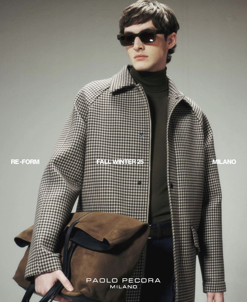 A brunette male model poses in a studio wearing a patterned overcoat, dark top and sunglasses, holding a brown suede bag in-hand.
